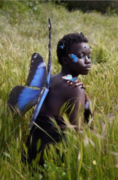 a young boy with blue butterflies on his body sits in tall grass and looks at the camera