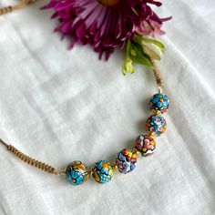 a necklace with colorful beads is on a white cloth next to a flower and a purple flower