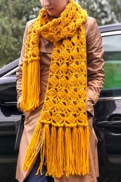 a woman standing next to a car wearing a yellow crocheted scarf with fringes