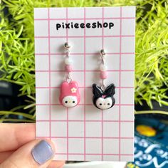 a pair of pink and white earrings with hello kitty on the front, sitting next to a plant