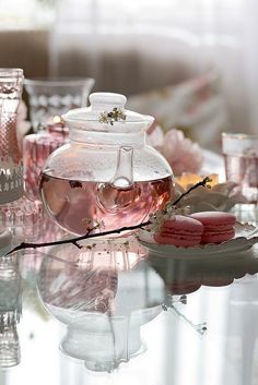 a glass table topped with pink macaroons and teapot filled with liquid sitting on top of it