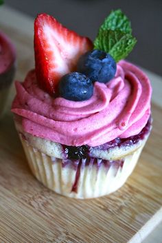 two cupcakes with pink frosting and blueberries on top are sitting on a cutting board