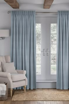 a living room with blue curtains and a white chair in front of a window that has wood beams on the ceiling