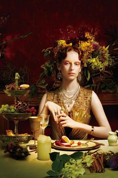 a woman sitting at a table with fruit and flowers in her hair, holding a glass of wine