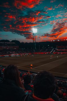 people watching a tennis match at night with the sun setting in the sky above them