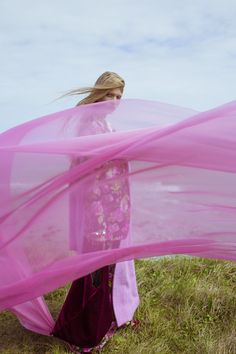 a woman is standing in the grass with her pink veil over her head and looking at the ground