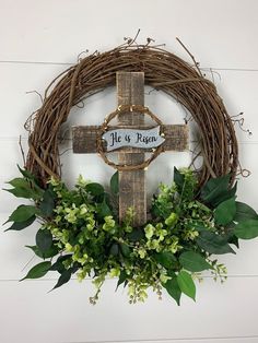 a cross with greenery is hanging on the wall next to a wreath that says, he is risen