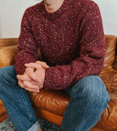 a man sitting on top of a brown leather chair wearing a red sweater and jeans