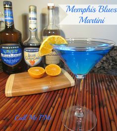 a blue martini with an orange slice on the rim next to bottles of alcohol and a cutting board