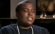 a man in a black shirt and pearls on his necklace is talking to the camera