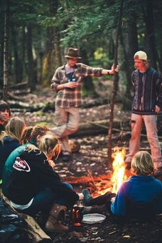 several people sitting around a campfire in the woods with one person pointing at something