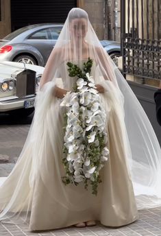 the bride is walking down the street in her wedding dress and veil with flowers on it