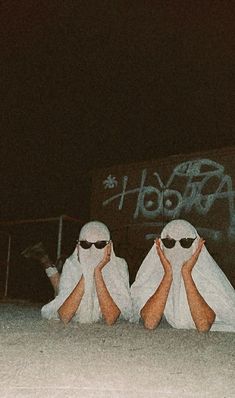 two people in white robes and sunglasses sitting on the ground with their hands up to their faces