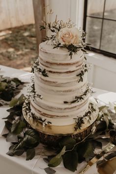 a white wedding cake with flowers and greenery on the table in front of a window