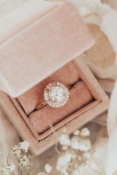 an engagement ring sits in a pink velvet box surrounded by white flowers and greenery