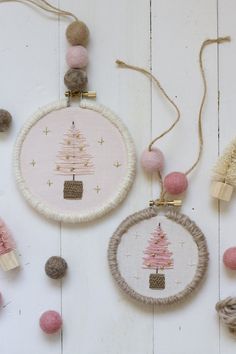 three hand embroidered ornaments hanging from strings on a white wooden surface with pink and grey pom poms