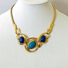 Vintage Kunio Matsumoto Era Trifari Modernist Necklace Gold Plated with Navy and Turquoise Acrylic Cabochons A great design that may or may not have been created by Kunio Matsumoto. It does not have his signature but it does have his style. It is marked Trifari. The necklace is composed of a center bib that is somewhat asymmetrical and set with a 1 inch by 11/16 of an inch larger turquoise color acrylic cabochon at the center. A smaller navy blue cabochon is set at each side. The smaller cabs measure 3/4 of an inch tall by 9/16 of an inch at the widest point. The centerpiece also features textured and highly polished elements. The necklace closes with a large textured hook, an element in the centerpiece design. Completing the necklace is a gorgeous bold flat snake or gas pipe chain 3/16 in Vintage Blue Jewelry With Unique Variations, Elegant Turquoise Necklace With Unique Variations, Modern Blue Oval Necklace, Formal Blue Cabochon Necklace, Elegant Turquoise Metal Necklace Gift, Elegant Turquoise Metal Necklace For Gift, Elegant Blue Cabochon Turquoise Necklace, Elegant Blue Turquoise Necklace With Cabochon, Elegant Turquoise Necklace As Gift