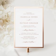 a white and gold wedding card on top of a table with flowers in the background