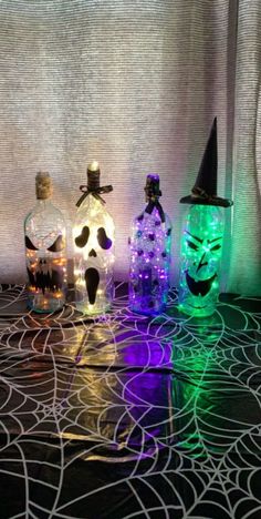 three lighted bottles with halloween decorations in them on a black tableclothed surface, one has a witch hat and the other is a skull