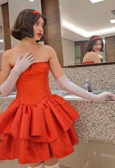 a woman in an orange dress and white gloves is looking at herself in the mirror