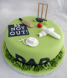 a cake decorated with green frosting and sports related items on the top, including a cricket bat, ball, tees, and helmet