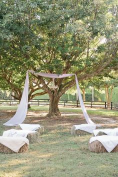 an outdoor wedding setup under a tree with white drapes and pillows on the ground