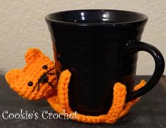 a crocheted orange cat sitting next to a black coffee cup