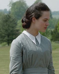 a woman standing in a field wearing a gray dress and white collared shirt with trees in the background