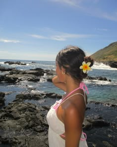 a woman with a flower in her hair standing on rocks near the ocean and looking at the water