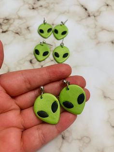 a person holding four green alien earrings in their hand, with the faces of an alien on them