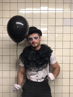 a man with makeup on his face standing next to a wall holding a black balloon