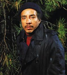 a man standing in front of a tree wearing a black coat and red shirt with his hands on his hips