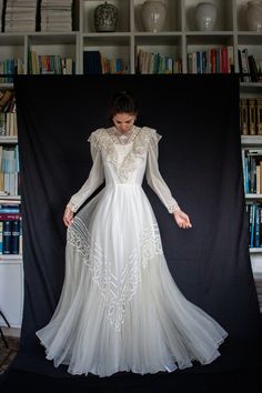 a woman is standing in front of a book shelf wearing a long white dress with sheer sleeves