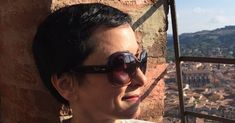 a woman wearing sunglasses standing next to a brick wall with a city in the background