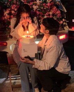 two people sitting in front of a cake with candles on it and one person covering his mouth