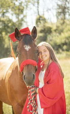 a girl in a red graduation gown standing next to a brown horse wearing a red sash