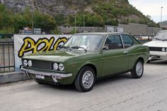 an old green car parked in front of a wall with graffiti on it