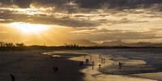 people are walking on the beach as the sun goes down in the distance with mountains in the background