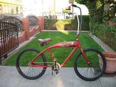 a red bicycle parked on the sidewalk in front of a fenced yard with potted plants