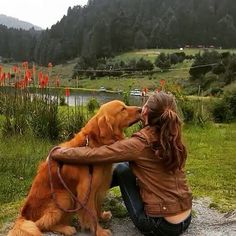 a woman sitting next to a dog on top of a grass covered field with mountains in the background