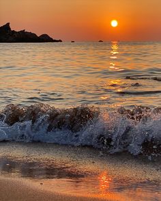 the sun is setting over the ocean with waves crashing in front of it and an island in the distance