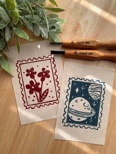 two stamps on top of a wooden table next to a plant and some markers with ink