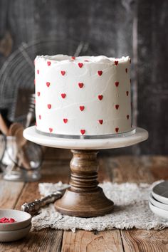 a white cake with red hearts on it sitting on top of a wooden table next to plates