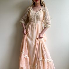 Oh so pretty: this peachy lace number looks darling! Vintage dress features a V-shaped neckline with long sleeves. Sleeves are composed of an ivory lace with 4 small pearl buttons at the cuffs. Small pearl trim along the collar. Lace up bodice. A line silhouette. Dress is lined with an ivory acetate fabric and a peachy pink cotton overlay. Original zipper in the back. Please note: there are some discoloration on the skirt. Please check photos provided. Medium weight. Label is Gunne Sax. Made in Silhouette Dress, Cotton Gowns, Gunne Sax Dress, 1970s Dresses, Gunne Sax, Peachy Pink, Pearl Buttons, Ivory Lace, Pink Cotton