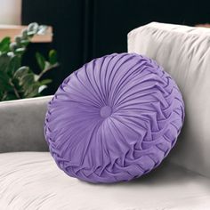 a round purple pillow sitting on top of a white couch next to a potted plant