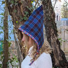 a woman wearing a blue and black plaid hoodie standing next to a tree in the woods