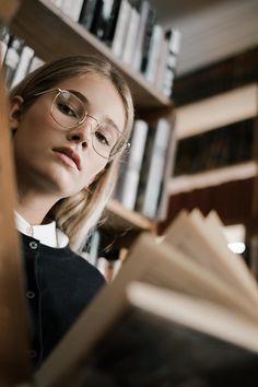 a woman wearing glasses reading a book in front of a bookshelf full of books