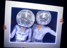 two people holding up a mirror with disco balls on their heads and hands behind them