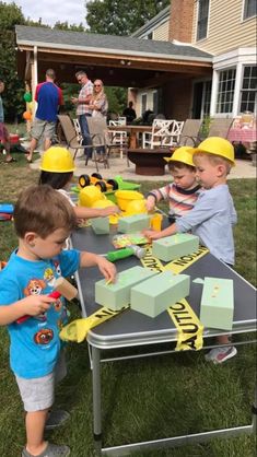 two young boys are playing with construction blocks on a table in the yard while others watch