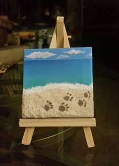 an easel with a painting on it and paw prints in the sand next to water